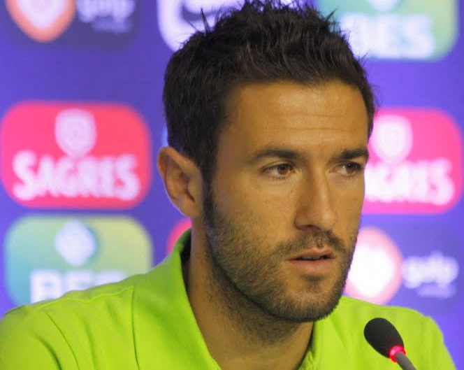 Hugo Viana at a press conference, wearing a bright green top with sponsor logos in the background